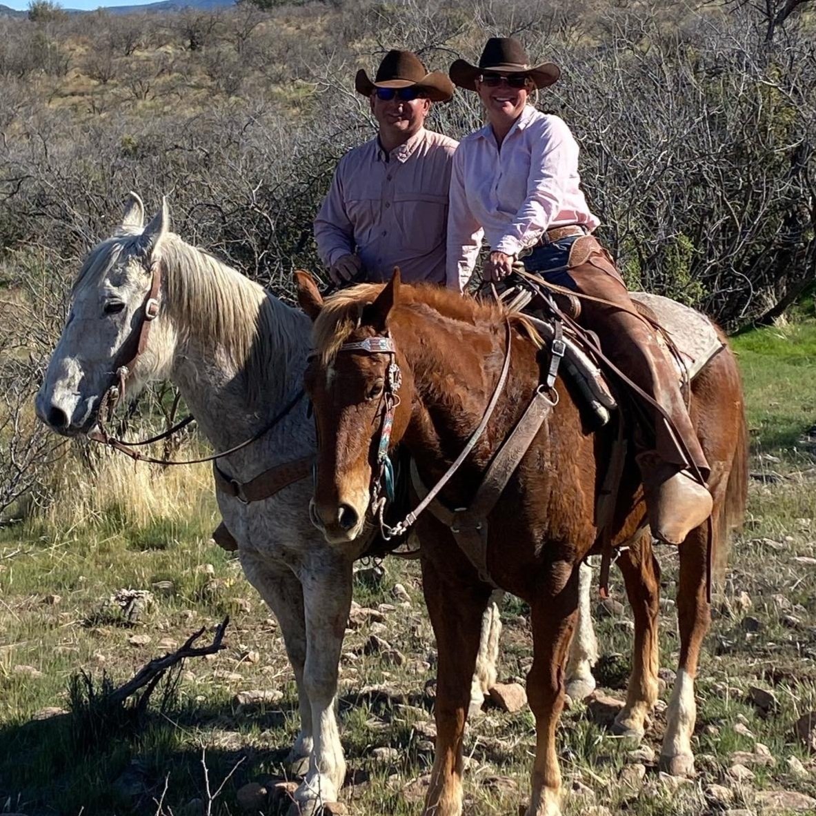 Two horseback riders, a man and a woman, wearing cowboy hats and western attire, pose on their horses in a natural outdoor setting with shrubs and hills in the background. The man is on a gray horse, while the woman rides a brown horse, showcasing a scenic equestrian experience.